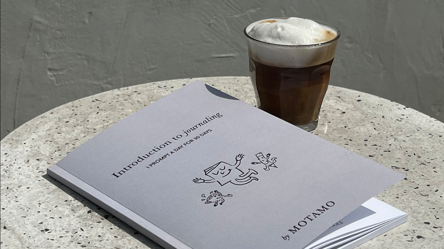 Minimalist picture of a notebook on a beige table next to a coffee with foam milk.